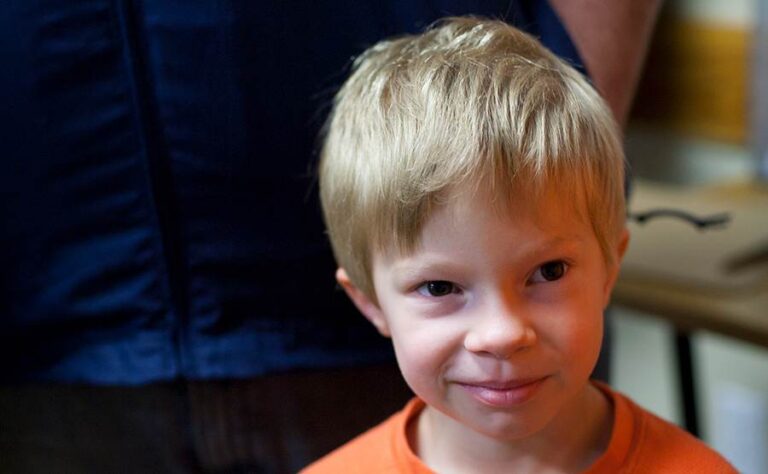 Young boy smiling indoors, wearing orange shirt.